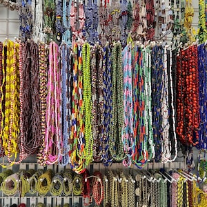 May include: A display of colorful beaded necklaces in various styles and lengths. The necklaces feature a wide array of colors, including red, blue, yellow, green, and pink. Some necklaces have intricate patterns, while others are solid colors.