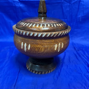 May include: A handcrafted wooden bowl with a lid. The bowl is brown with white accents and a dark brown base. The lid has a small, conical handle. The bowl is likely used for storage or decoration.
