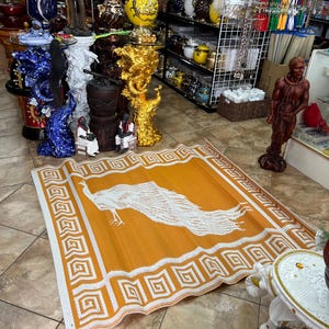 May include: An orange and white rectangular mat with a peacock design. The mat has a white border with a repeating geometric pattern. The peacock is depicted in white, with its tail feathers spread across the mat.