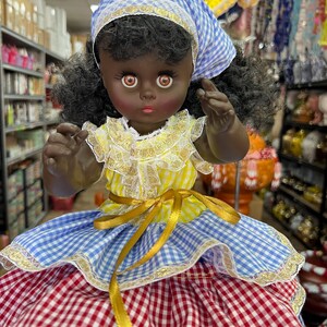 May include: A vintage doll with dark skin, wearing a yellow and white top with lace trim, a blue and white gingham headscarf, and a red and white gingham skirt. The doll has large brown eyes and is posed with its arms raised.