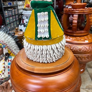 May include: A conical hat featuring a green and yellow fabric exterior adorned with rows of white cowrie shells. The base is densely covered in a thick layer of hanging cowrie shells, while a vertical panel on the side displays additional shells.