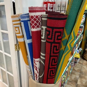 May include: A variety of rolled-up woven mats in a cardboard box. The mats display a range of colors and patterns, including geometric designs in red, blue, yellow, and green. The mats are displayed in a retail setting.