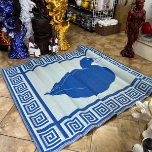 May include: A rectangular blue and white mat with a large blue leaf design in the center. The mat has a border with a repeating geometric pattern. The mat is made of a woven material and is displayed on a tiled floor.
