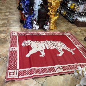 May include: A red and white rectangular mat featuring a white tiger design. The mat has a white geometric border. The mat is displayed on a tiled floor in a store setting, with various decorative items in the background.