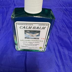 May include: A square glass bottle of scented oil with a white cap. The label reads "CALM BALM" and "BÁLSAMO TRANQUILO". The bottle contains 1 fl oz of oil. The background is a solid blue color.