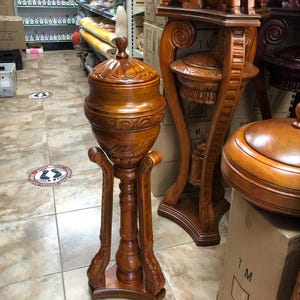 May include: A tall, ornate wooden urn with a lid, set on a decorative stand. The urn and stand are carved from a warm-toned wood, featuring intricate patterns and designs. The stand has a flared base and curved supports. Other wooden decorative items are visible in the background.