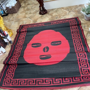 May include: Black and red rectangular mat with a large red face design in the center. The mat has a black background with a red Greek key border. The face has eye and mouth cutouts.