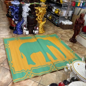 May include: A rectangular, woven mat with a large green elephant silhouette on a yellow background. The mat has a green border with a geometric design. The mat is displayed on a tiled floor in a shop setting.