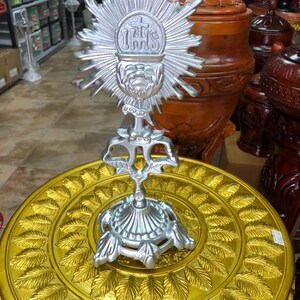 May include: A silver-colored monstrance with a sunburst design, featuring the letters IHS and a cross. It is displayed on a gold-colored, leaf-patterned stand. The monstrance is a religious item.