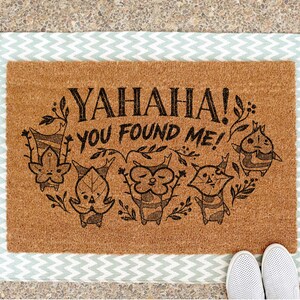 K&ouml;nnte beinhalten: Eine rechteckige Fu&szlig;matte mit einer braunen Kokosoberfl&auml;che und einem hellblau-wei&szlig;en Zickzack-Rand. Die Matte zeigt den Text "YAHANA! YOU FOUND ME!" mit Cartoon-Figuren.