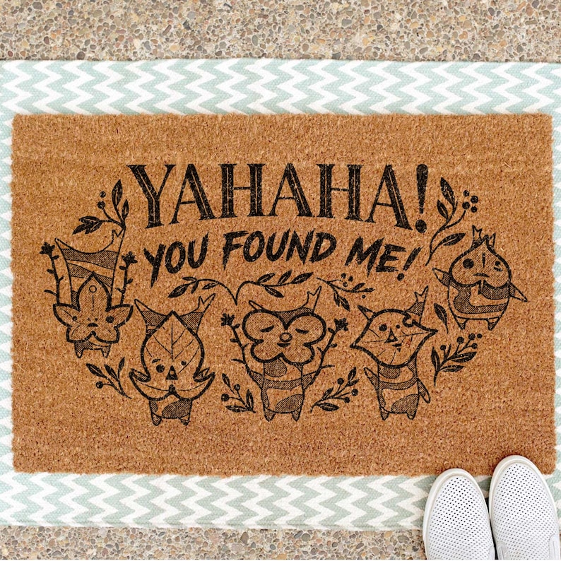 K&ouml;nnte beinhalten: Eine rechteckige Fu&szlig;matte mit einer braunen Kokosoberfl&auml;che und einem hellblau-wei&szlig;en Zickzack-Rand. Die Matte zeigt den Text "YAHANA! YOU FOUND ME!" mit Cartoon-Figuren.