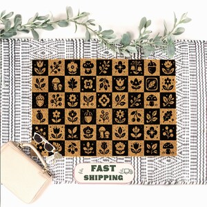 May include: A rectangular doormat featuring a checkerboard pattern of alternating natural tan coir and black squares. Each square contains a unique botanical illustration, including various flowers, mushrooms, acorns, and leaves in a bold graphic style.