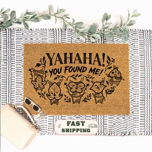 K&ouml;nnte beinhalten: Rechteckige Fu&szlig;matte aus Naturkokosfaser. Der Schriftzug "YAHAHA! YOU FOUND ME!" ist in Schwarz aufgedruckt, zusammen mit einer Grafik von f&uuml;nf Cartoon-Figuren. Die Matte liegt auf einem schwarz-wei&szlig; gemusterten Teppich, mit Gr&uuml;nzeug.
