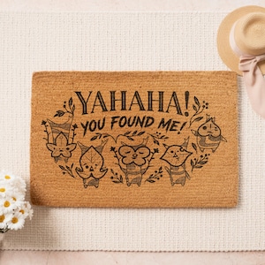 K&ouml;nnte beinhalten: Eine rechteckige Kokos-Fu&szlig;matte mit dem schwarzen Text "YAHАHA! YOU FOUND ME!". Das Design umfasst cartoonartige Figuren und florale Elemente. Ein Strohhut mit rosa Band ist im Hintergrund zu sehen.