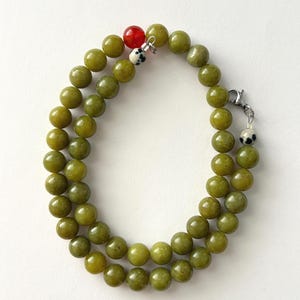 May include: A double-strand necklace featuring smooth, round olive green beads. The necklace includes a single bright red bead, two small white beads with black spots, and a silver-toned metal lobster claw clasp closure.