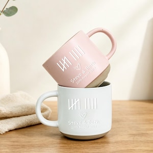 May include: Two ceramic mugs stacked vertically, one light pink and one white. Each mug features a tally mark design, a small heart icon, and the engraved text "Steve & Beth 20th August 2017". The mugs have a smooth finish with a contrasting base.