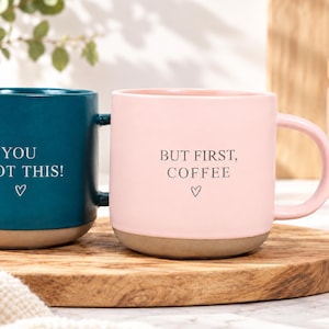 Könnte beinhalten: Ein dunkelblauer Becher mit dem Text YOU GOT THIS! und einem Herzsymbol sowie ein hellrosa Becher mit dem Text BUT FIRST, COFFEE und einem Herzsymbol. Beide Keramikbecher haben einen glatten farbigen oberen Teil und einen naturfarbenen Boden.