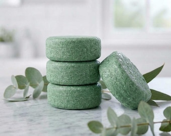 May include: A stack of three round, sage green solid shampoo bars with a textured, granular surface, accompanied by a fourth matching bar leaning against the stack, all surrounded by sprigs of green eucalyptus leaves.