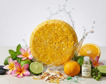 May include: A round yellow shampoo bar surrounded by water splashes, pink plumeria flowers, lime slices, a whole orange, coconut flakes, green grapes, and a small glass bottle labeled Citrus Mint Bliss Scentuales.