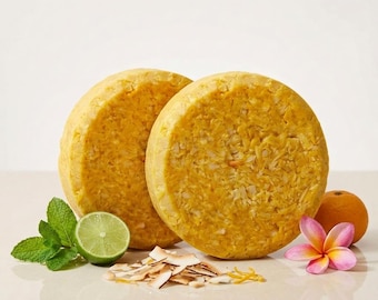 May include: Two round, textured, golden-yellow shampoo bars are displayed alongside a halved lime, fresh green mint leaves, a small pile of dried coconut flakes, a whole orange, and a single pink and yellow plumeria flower blossom.