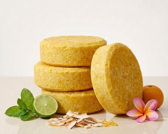 May include: Three stacked round yellow shampoo bars next to one upright yellow shampoo bar, a sliced lime, fresh mint leaves, scattered coconut flakes, a whole orange, and a pink and yellow plumeria flower on a smooth surface.