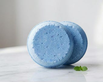 May include: Two round, light blue bath bombs with a textured, granular surface. The bath bombs are stacked slightly offset from one another, and a single small green mint leaf is placed near the base of the items.