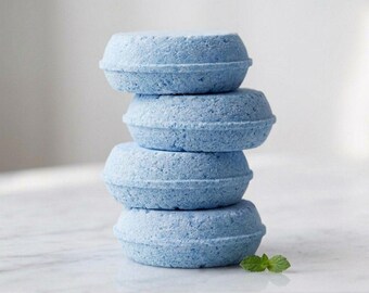 May include: A stack of four round, light blue solid shampoo bars with a textured, porous surface. The bars are cylindrical with a slightly tapered edge. A single small green mint leaf is placed on the surface next to the base of the stack.
