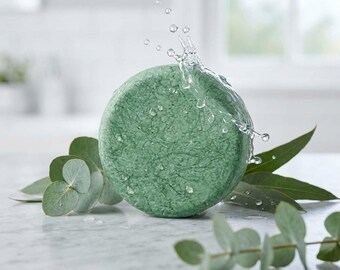 May include: A round, textured green shampoo bar with water splashing against its surface. The bar is positioned behind and among several green eucalyptus leaves, with visible water droplets resting on the leaves and the bar itself.