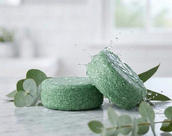May include: Two round, light green shampoo bars with a textured surface. One bar lies flat while the other is tilted, with water droplets splashing off its surface. Several green eucalyptus leaves are arranged around the bars.