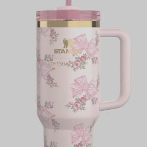 May include: A light pink Stanley tumbler with a floral and bow pattern, featuring a gold-tone rim, a clear lid, and a matching pink straw topped with a bow. The text STANLEY and LOVESHACKFANCY is printed in gold on the front of the insulated cup.
