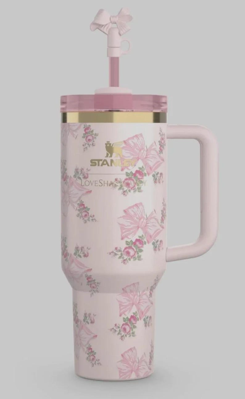 May include: A light pink Stanley tumbler with a floral and bow pattern, featuring a gold-tone rim, a clear lid, and a matching pink straw topped with a bow. The text STANLEY and LOVESHACKFANCY is printed in gold on the front of the insulated cup.