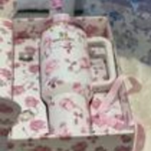 May include: A white ceramic mug with a pink floral pattern and a matching pink floral gift box. The mug features a handle and a pink lid topper. The box interior and exterior are lined with the same pink floral print, and a pink ribbon is attached.