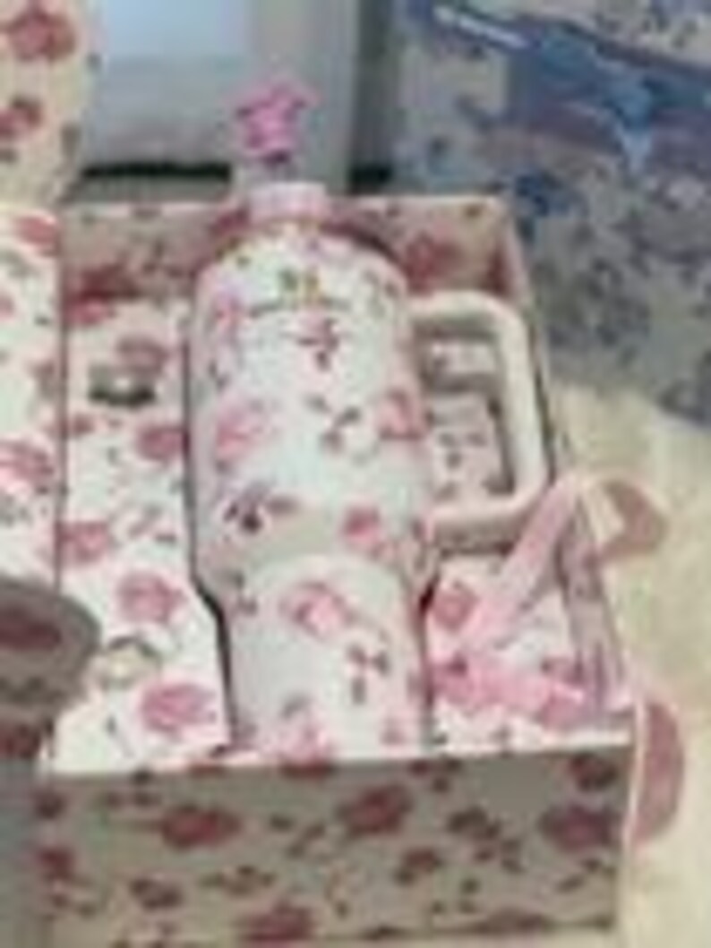 May include: A white ceramic mug with a pink floral pattern and a matching pink floral gift box. The mug features a handle and a pink lid topper. The box interior and exterior are lined with the same pink floral print, and a pink ribbon is attached.