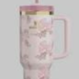 May include: A light pink insulated tumbler with a handle, featuring a repeating pattern of small pink bows. The tumbler includes a matching pink lid with a gold-colored rim and a clear straw. The word Stanley is printed in small letters on the front.