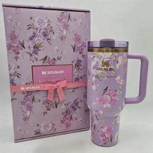 May include: A lavender Stanley tumbler with a floral pattern and a matching box. The tumbler features a handle, a clear lid, and the Stanley LoveShackFancy logo. The box is decorated with the same floral print and includes a pink ribbon detail.