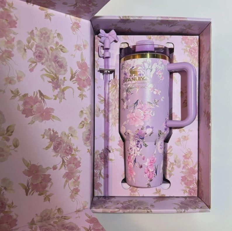 May include: A lavender Stanley tumbler with a floral pattern, a matching handle, and a clear lid. The tumbler includes a long, clear straw topped with a small lavender bow accessory. The set is presented in a matching floral-patterned box.
