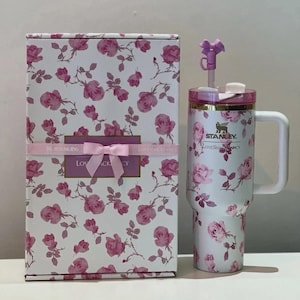 May include: A white Stanley tumbler with a pink rose floral pattern, a white handle, a pink lid, and a purple bow straw topper. Next to it is a matching rectangular box featuring the same floral print, a pink ribbon, and the text Stanley LoveShackFancy.