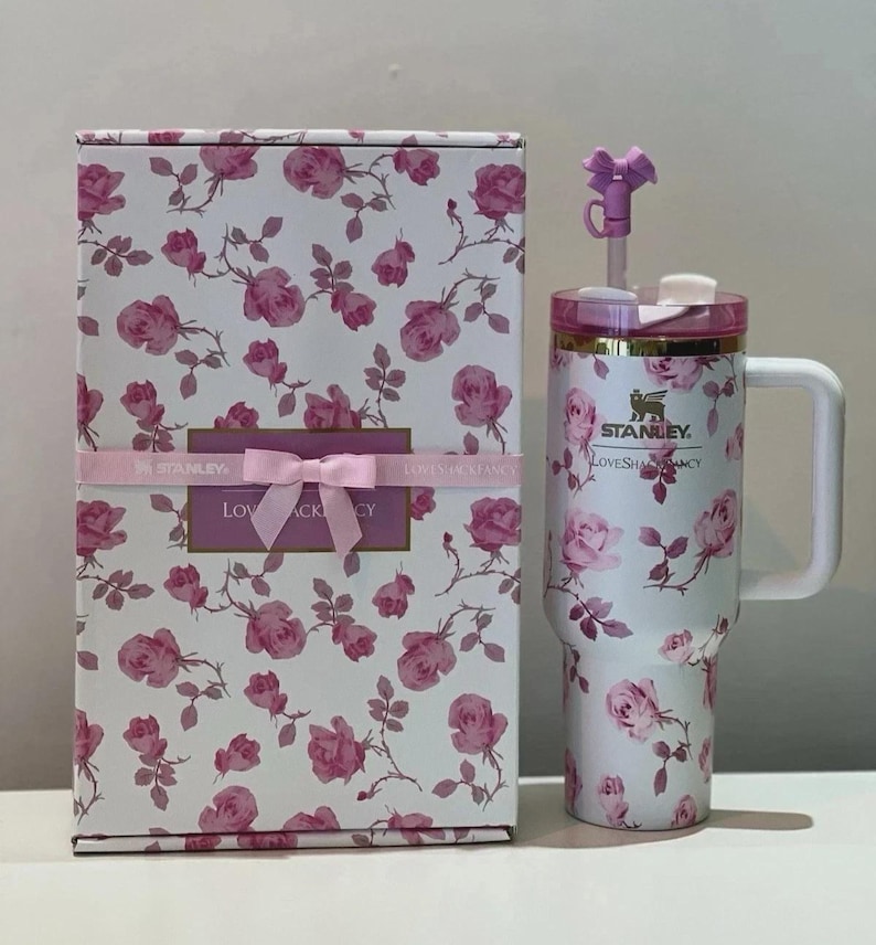 May include: A white Stanley tumbler with a pink rose floral pattern, a white handle, a pink lid, and a purple bow straw topper. Next to it is a matching rectangular box featuring the same floral print, a pink ribbon, and the text Stanley LoveShackFancy.