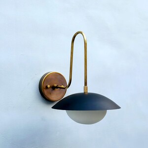 May include: A contemporary wall light featuring a black lampshade and a brass-toned curved arm. The light has a circular, bronze-coloured base and a frosted glass globe. The design is minimalist and stylish, suitable for interior lighting.