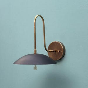 May include: A wall-mounted lamp featuring a dark blue, semi-circular shade and a brass-coloured curved arm. The lamp has a round, brass-coloured base and a visible lightbulb. The design is minimalist and modern.
