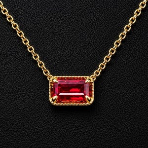 Puede incluir: Un collar de cadena dorada con un colgante de piedra preciosa roja de corte esmeralda rectangular. La piedra está engastada en un marco dorado con borde de cuentas y cuatro garras, suspendido de una delicada cadena de eslabones dorados.
