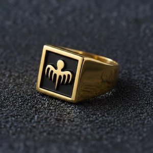 May include: A gold-toned signet ring featuring a square face with a matte black inlay. Centered on the black square is a raised, gold-toned emblem depicting a stylised bird or winged creature with a rounded head and five distinct downward feathers.