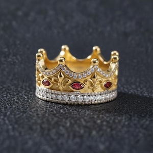 Könnte beinhalten: Ein goldener Kronenring mit einem Band aus kleinen klaren Steinen an der Basis, einer Reihe roter Steine im Marquise-Schliff und Fleur-de-Lis-Akzenten. Der obere Rand des Rings ist gewellt und mit winzigen klaren Steinen besetzt.