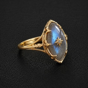 Könnte beinhalten: Ein goldener Ring mit einem länglichen, marquiseförmigen Labradorit-Edelstein mit blauem, irisierendem Schimmer. Die Fassung enthält einen kleinen, sternförmigen Akzent mit einem winzigen Stein und zarte blattartige Gravuren auf der Ringschiene.