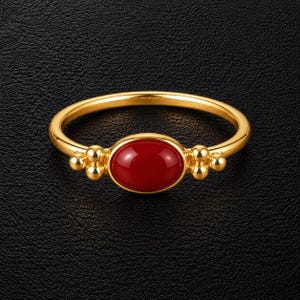 May include: A gold ring featuring a smooth, oval-shaped red stone set in the center. The band is accented on both sides of the stone with a cluster of three small, polished gold spheres arranged in a vertical formation, creating a balanced design.