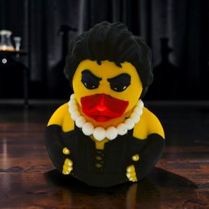 Könnte beinhalten: Eine gelbe Quietscheente mit schwarzem Haar, dunklem Lidschatten und einem roten Schnabel. Sie trägt eine weiße Perlenkette und ein schwarzes Korsett-Outfit mit passenden schwarzen Handschuhen. Die Figur weist eine texturierte Oberfläche auf.