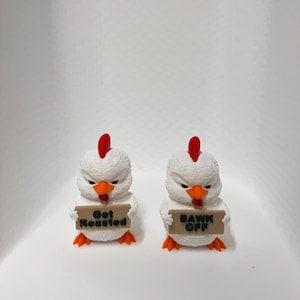 Pode incluir: Duas figuras de galinha brancas com cristas vermelhas e bicos e pés laranja. Cada uma segura uma placa marrom com texto preto. Uma diz "Get Roasted" e a outra "BAWK OFF".