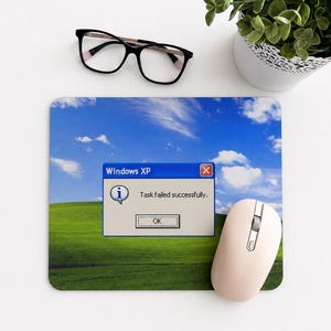 May include: A rectangular mousepad featuring the iconic Windows XP "Bliss" background with a blue sky and green hills. The mousepad displays a Windows XP error message: "Task failed successfully." A pink computer mouse and black eyeglasses are also present.