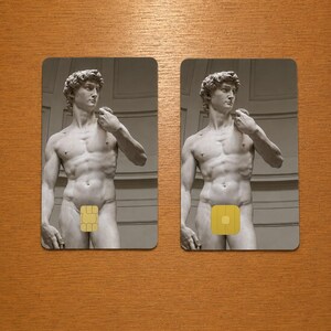 Op de afbeelding: Twee rechthoekige kaarten met een grijswaardenafbeelding van Michelangelo's David-standbeeld. Elke kaart heeft een gele vierkant die de geslachtsdelen van het standbeeld bedekt. De kaarten liggen op een houten oppervlak.