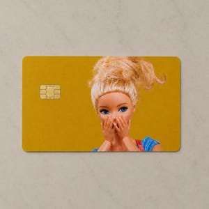 Pu&ograve; includere: Una carta di credito rettangolare con uno sfondo giallo senape solido e un chip metallico dorato sul lato sinistro. Il lato destro mostra una bambola con capelli biondi raccolti in uno chignon spettinato, occhi blu e mani sulla bocca.
